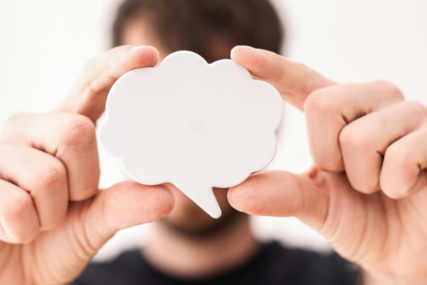 Eine Person mit verschwommenem Gesicht hält eine weiße, wolkenförmige Sprechblase vor sich. Die Sprechblase ist scharf und verdeckt die Gesichtszüge der Person. Der Hintergrund ist schlicht weiß.