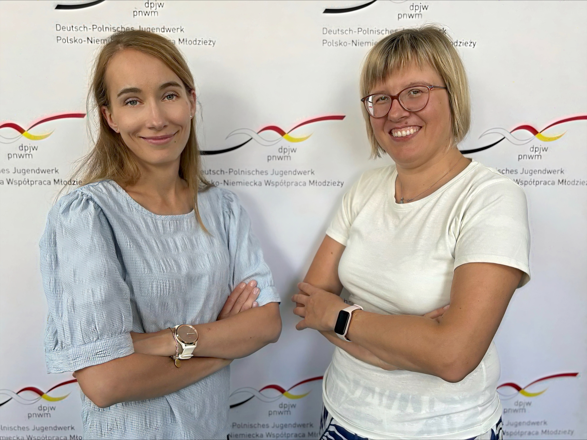 Zwei Frauen stehen nebeneinander, lächelnd und mit verschränkten Armen vor einem Hintergrund mit dem Logo und dem Text des Deutsch-Polnischen Jugendwerks (DPJW/PNWM). Beide tragen Brillen und legere Oberteile.