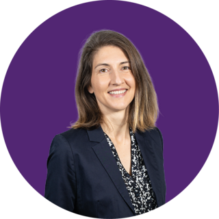 Eine Frau mit schulterlangem braunem Haar, die einen dunklen Blazer über einer gemusterten Bluse trägt, lächelt in die Kamera. Sie steht vor einem einfarbig violetten, runden Hintergrund.