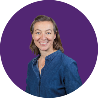 Eine Frau mit schulterlangem braunem Haar und blauer Bluse lächelt in die Kamera. Der Hintergrund ist ein einfarbiger lila Kreis.