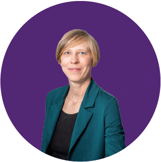 Vor einem einfarbigen, violetten, runden Hintergrund ist eine Person mit kurzen blonden Haaren zu sehen, die einen blaugrünen Blazer und ein schwarzes Oberteil trägt und lächelt – perfekt für eine „Über uns“-Vorstellung.