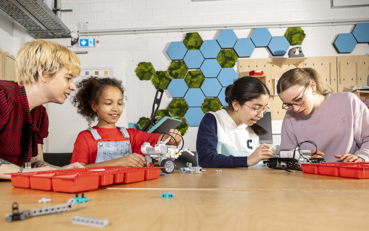 Drei Schülerinnen und eine Lehrkraft sitzen verteilt um einen Tisch und arbeiten in Zweiergruppen daran, Roboter zusammenzubauen. Die Lehrkraft schaut auf der linken Seite gespannt zu. Vor ihnen stehen zwei rote Aufbewahrungsboxen mit Bauteilen. Im Hintergrund sind ein Holzschrank und kleine Akustik-Wandpaneele aus blauen und grünen Hexagonen zu sehen. Die Schülerinnen sind etwa 12 bis 14 Jahre alt. Das Bild ist waagerecht aufgenommen.